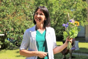 Sieviete ar ziediem rokās, ģērbusies baltā mētelī, stāv dārzā pie Rīgas 1. slimnīcas.