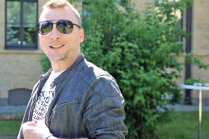 Vīrietis ar saulesbrillēm un džinsu jaku, stāv dārzā pie Rīgas 1. slimnīcas. Fons ar zaļumiem.