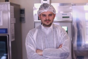 Vīrietis medicīniskajā apģērbā stāv virtuvē, rokas saliktas krūtīs, aiz viņa redzami ledusskapji.