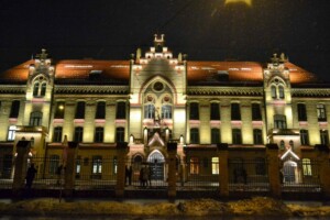Rīgas 1. slimnīcas ēka naktī, apgaismota ar siltu gaismu, sniegots fons un cilvēki pie ieejas.