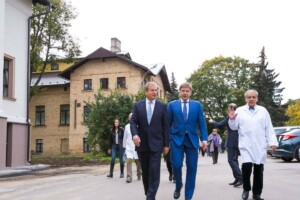 Divi vīrieši uz ielas, viens tērpies uzvalkā, otrs baltā mētelī, fonā Rīgas 1. slimnīcas ēka.