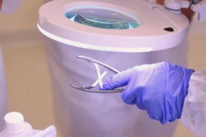 Rokas ar zilu cimdu tur medicīniskos instrumentus zem palielināmā stikla. Rīgas 1. slimnīca.