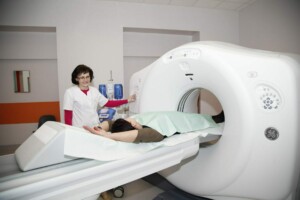 Pacients guļ uz izmeklēšanas galda, blakus stāv medicīnas darbinieks un CT skeneris.