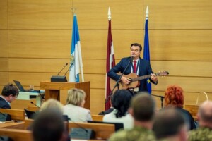 Vīrietis ar ģitāru uzstājas telpā ar Latvijas un Eiropas Savienības karogiem. Publikumā redzami cilvēki.
