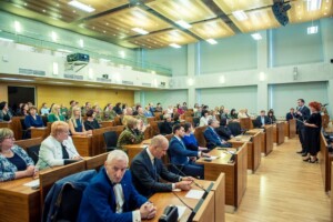 Sanāksme Rīgas 1. slimnīcā, dalībnieki sēž zālē, runātāji uz skatuves.