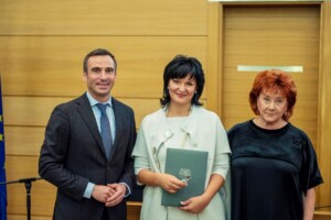 Trīs cilvēki stāv kopā, turēdami dokumentu, fons ar koka apdari un Latvijas karogu. Rīgas 1. slimnīca.
