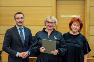 Trīs cilvēki stāv telpā, tērpušies melnā, tur rokās dokumentus. Rīgas 1. slimnīcas pasākums.