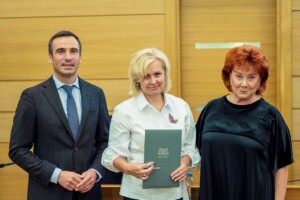 Trīs cilvēki stāv pie galda, centrā sieviete ar dokumentu "Rīgas dome". Fons ar koka paneļiem.