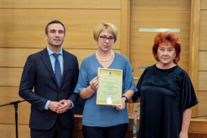 Divi cilvēki stāv blakus sievietei, kas tur diplomu ar tekstu "Rīgas 1. slimnīca". Fons koka paneļi.