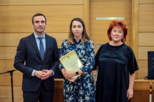 Trīs cilvēki stāv zālē, vidū ir sieviete ar diplomu "Pateicības raksts" Rīgas 1. slimnīcā.