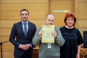 Divi cilvēki stāv blakus vīrietim, kurš tur pateicības rakstu ar tekstu "Rīgas 1. slimnīca".
