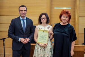 Trīs cilvēki stāv pie galda, vidū sieviete ar sertifikātu "Pateicības raksts" no Rīgas 1. slimnīcas.