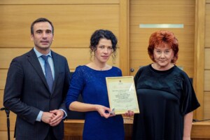 Divi cilvēki stāv blakus sievietei, kas tur pateicības rakstu ar tekstu "Pateicības raksts". Rīgas 1. slimnīca.