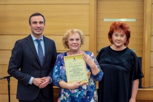 Divi cilvēki stāv blakus sievietei, kas tur sertifikātu ar tekstu "Pateicības raksts". Rīgas 1. slimnīca.