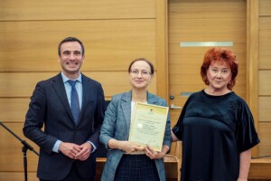 Trīs cilvēki stāv pie koka sienas, vidū sieviete ar diplomu "Paieciības raksts".