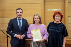 Trīs cilvēki stāv pie galda, jaunā sieviete tur diploma sertifikātu ar tekstu "Pateicības raksts".
