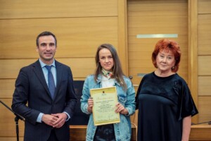 Trīs cilvēki stāv pie galda, vidū sieviete ar pateicības rakstu Rīgas 1. slimnīcai par ieguldījumu.