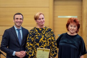 Trīs cilvēki stāv pie koka sienas, vidū sieviete ar sertifikātu "Pateicības raksts". Rīgas 1. slimnīca.