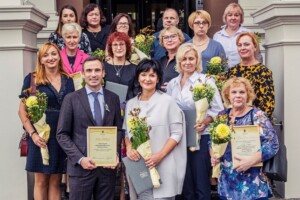 Grupa cilvēku ar ziediem un diplomiem, stāvot pie Rīgas 1. slimnīcas ieejas. Redzami vārdi "Paldies" un "Apsveicam".