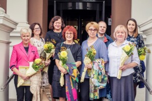 Grupa cilvēku ar ziediem, stāv pie Rīgas 1. slimnīcas ieejas, svinot notikumu. Redzama pateicības balva.