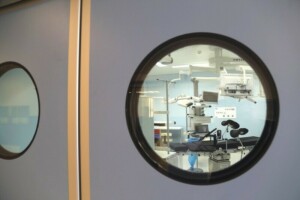 Skats uz operāciju zāli caur apaļu logu, redzama operāciju gulta un medicīnas iekārtas.