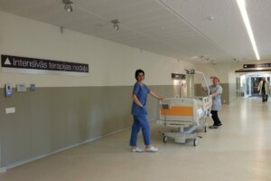 Divi mediķi intensīvās terapijas nodaļā, pārvietojot gultu. Redzams uzraksts "Intensīvās terapijas nodaļa".