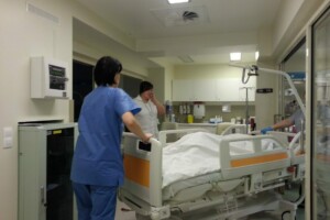 Divas medicīnas darbinieces strādā slimnīcas telpā pie gultas ar pacientu. Redzama medicīnas iekārta.