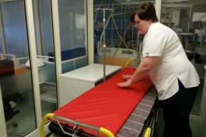 Māsa izlīdzina sarkano gultas pārklāju slimnīcas telpā. Redzama gulta un medicīnas aprīkojums.
