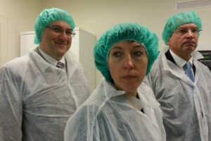 Trīs cilvēki medicīniskās drēbēs un zaļās cepurēs, stāvot Rīgas 1. slimnīcas telpās.