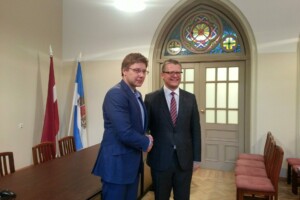 Divi vīrieši satiekas un pasniedz roku, fons ar Latvijas karogu un krāsainu logu. Rīgas 1. slimnīca.