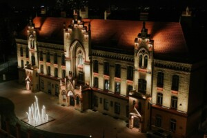 Rīgas 1. slimnīcas ēka apgaismota naktī, redzama strūklaka priekšā.