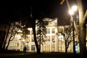 Rīgas 1. slimnīca naktī, apgaismota, ar kokiem un gaismas stabiem priekšplānā.