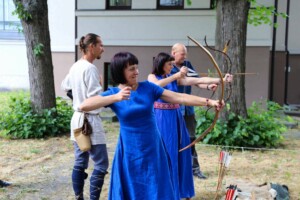 Cilvēki šauj ar loku, tērpti tradicionālās drēbēs, Rīgas 1. slimnīcas pagalmā.