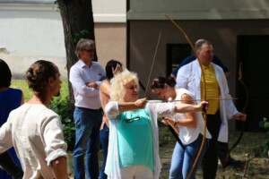 Cilvēki šauj ar loku ārā, Rīgas 1. slimnīcas pasākumā, daži skatās, citi piedalās.