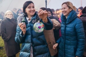 Sieviete ar sarkanu sirdi rotātu Ziemassvētku rotājumu, apkārt citi cilvēki svinīgā pasākumā.