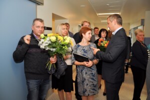 Grupa cilvēku Rīgas 1. slimnīcā, daži ar ziediem, sarunājas un sveic viens otru.