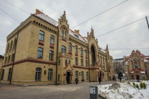 Rīgas 1. slimnīcas ēka ar vēsturisku arhitektūru un sniega segumu uz jumta. Redzami gājēji un apkārtne.