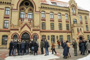 Rīgas 1. slimnīcas ēka ar cilvēku grupu pie ieejas un žurnālistiem fonā. Ziemas ainava ar sniegu.
