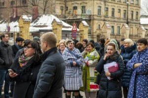 Cilvēki gaida pie Rīgas 1. slimnīcas, tērpušies ziemas apģērbā, daži ar dokumentiem rokās.
