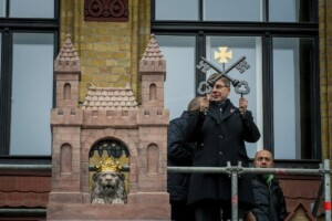 Vīrietis ar atslēgām stāv pie ēkas, kurā redzams lauvas statuja un logi. Rīgas 1. slimnīca fons.
