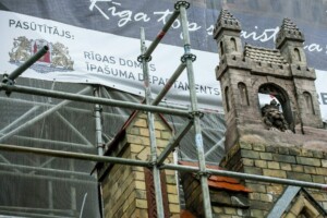 Attēlā redzama būvniecības skatuve ar Rīgas Domes Īpašuma departamenta reklāmu un dekoratīvu torni.