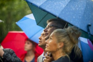 Cilvēki ar lietussargiem skatās debesīs, lietus pilieni krīt uz viņu sejām. Rīgas 1. slimnīca.