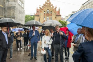 Grupa cilvēku ar lietussargiem stāv pie vēsturiskas ēkas, notiek diskusija par Rīgas 1. slimnīcas projektu.