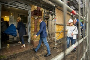 Cilvēki ieiet Rīgas 1. slimnīcas ēkā, lietus laikā, redzami būvniecības darbi. Uz durvīm norādes par reģistratūru.