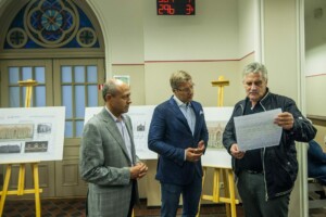 Trīs vīrieši apspriež plānus pie stenda ar arhitektūras projektiem Rīgas 1. slimnīcai. Fons ar logu un pulksteni.