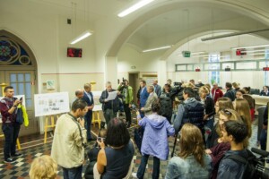 Pasākums Rīgas 1. slimnīcā, cilvēki pulcējušies, fiksēti plāni un runājošs vīrietis. Redzami žurnālisti un kameras.