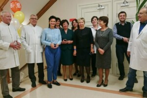 Rīgas 1. slimnīcas darbinieki svin jubileju, stāvot kopā pie kāpnēm ar baloniem un smaidiem.