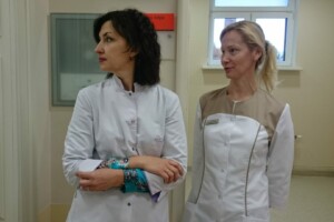 Divas sievietes medicīnas tērpos stāv slimnīcas telpā, skatoties uz sāniem. Redzama zīme "Pacientu telpa".