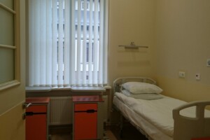 Gulta slimnīcas istabā ar logiem, aiz kuriem redzami aizkari un divi skapīši blakus.
