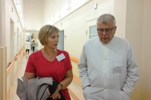 Divi cilvēki sarunājas slimnīcas koridorā. Sieviete valkā sarkanu kreklu, vīrietis - baltu halātu.